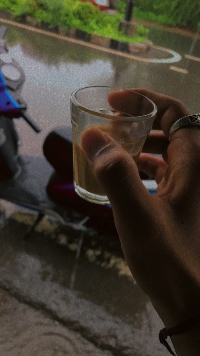 Eine Person mit einem Bierglas vor einem Fenster, daneben ein Motorroller und im Hintergrund Bäume, Pfähle und eine Straße.