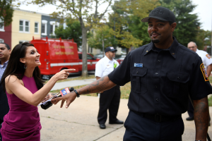 Eine Frau in einem lilafarbenen Kleid spricht mit einem Polizeibeamten in einem schwarzen Kleid und einer Mütze, mit mehreren Personen, Fahrzeugen, Bäumen, Gebäuden und einem weißen Himmel im Hintergrund.