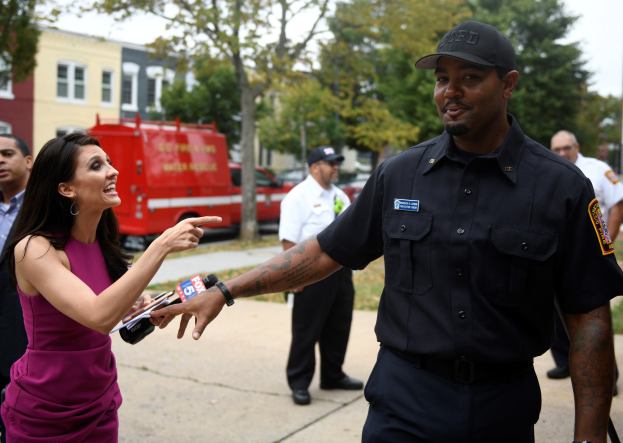 Eine Frau in einem lilafarbenen Kleid spricht mit einem Polizeibeamten in einem schwarzen Kleid und einer Mütze, mit mehreren Personen, Fahrzeugen, Bäumen, Gebäuden und einem weißen Himmel im Hintergrund.