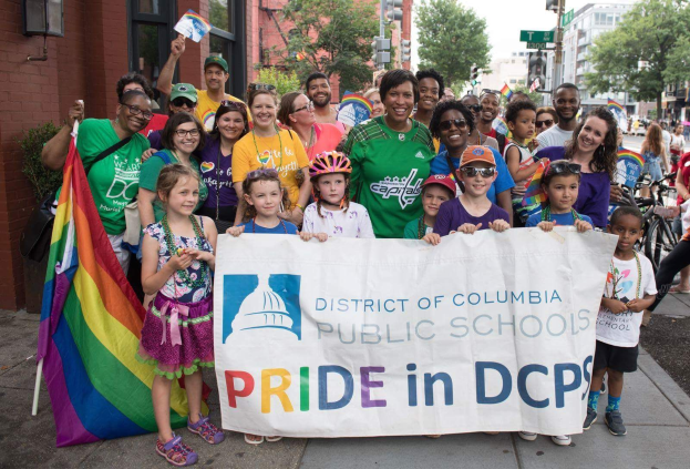 Gruppe von Menschen auf dem Boden, einige halten ein Banner mit der Aufschrift "District of Columbia Public Schools Pride in DCPS", mit Kindern, die eine Fahne halten, einer Person auf dem Fahrrad, Bäumen, Gebäuden, Straßen-Elementen und einem bewölkten Himmel.