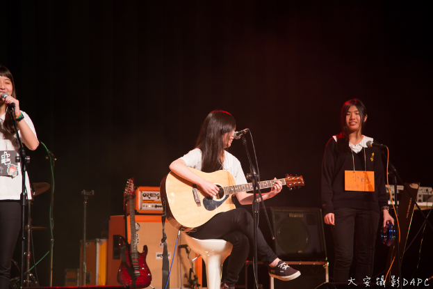 Drei Mädchen auf der Bühne, zwei singen in Mikrofone und eine hält ein Musikinstrument, mit einer Gitarre, einem Lautsprecher und einer Schalttafel zu sehen.