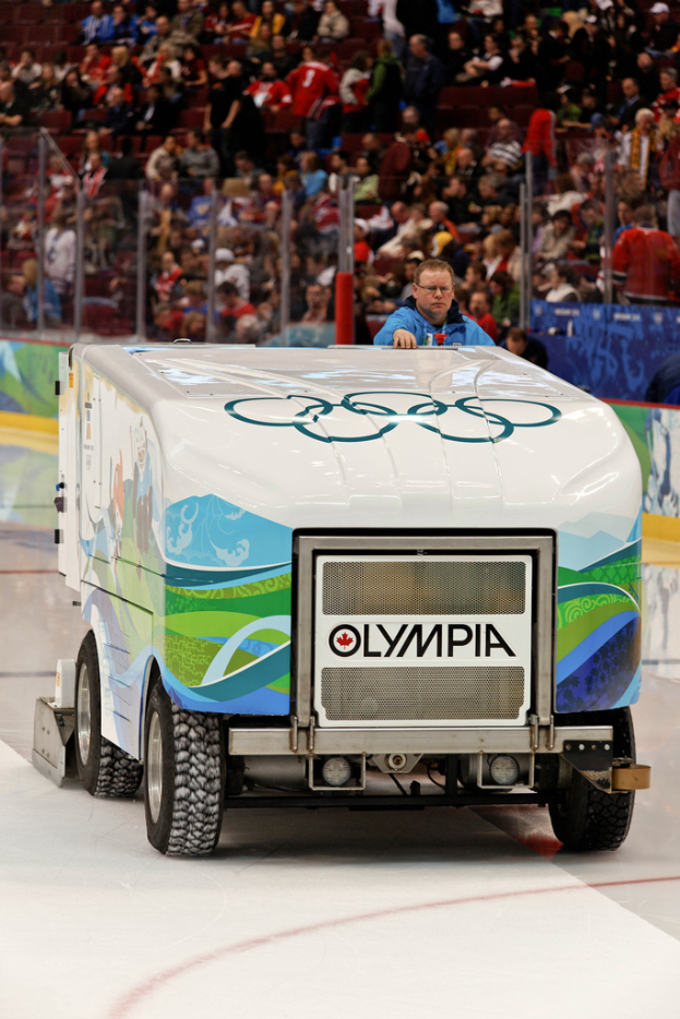 Ein Mann in einer blauen Jacke und Brille steht auf einem Fahrzeug, mit einem Publikum, das darüber sitzt, und einem Zaun und einem Werbeplakat im Hintergrund.