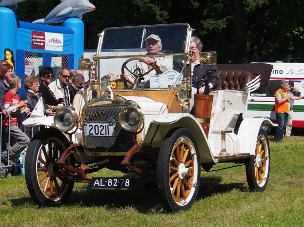 Ein Mann fährt ein Oldtimer-Auto an einer Menge vorbei, die Kappen trägt und auf einer Rasenfläche mit einem Geländer, einem aufblasbaren Objekt, Bäumen und anderen Fahrzeugen im Hintergrund steht.