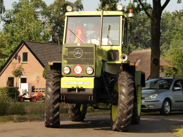 Grüner Traktor auf einer Straße neben einem Auto mit einem sichtbaren Fahrer, Häuser und Bäume im Hintergrund unter einem klaren blauen Himmel.
