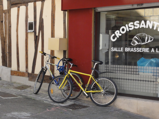Zwei Fahrr├Ąder nebeneinander auf einem Gehweg vor einem Laden mit einer Glaswand und Text, mit einem Objekt an der Wand links von den Fahrr├Ądern.