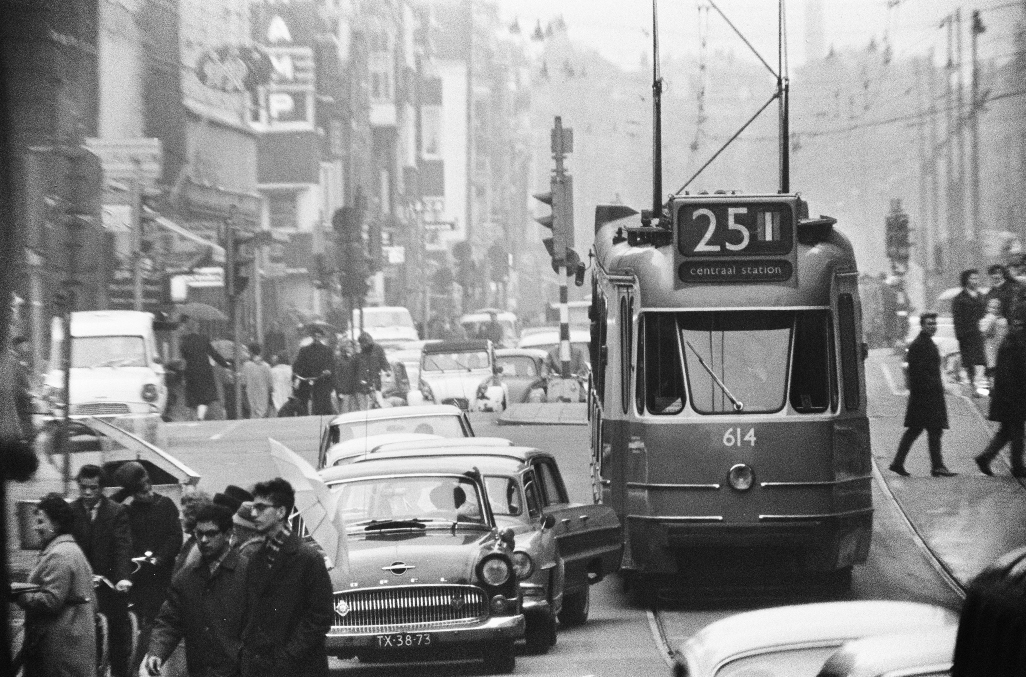 Schwarz-weiß-Foto einer belebten Stadtstraße mit Fahrzeugen einschließlich einem Bus, Auto und Lieferwagen, Fußgängern auf dem Gehweg, einige halten Regenschirme, und Gebäuden, Pfählen und Verkehrszeichen im Hintergrund.