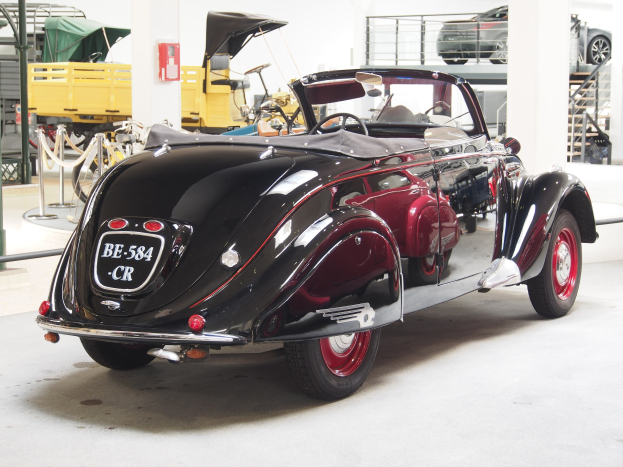 Schwarzes und rotes Auto in einer Garage mit anderen Fahrzeugen, Pfählen, Ketten und Gegenständen im Hintergrund geparkt, mit einem Feuerlöscher an der Wand montiert.