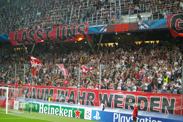 Ein Fußballspiel in einem Stadion mit einer großen Zuschauermenge, saftig grünem Rasen, einem Torpfosten links, Bodenbanner, Menschen mit Fahnen und Hintergrundgeländer, Tafeln, Lampen und andere Gegenstände.
