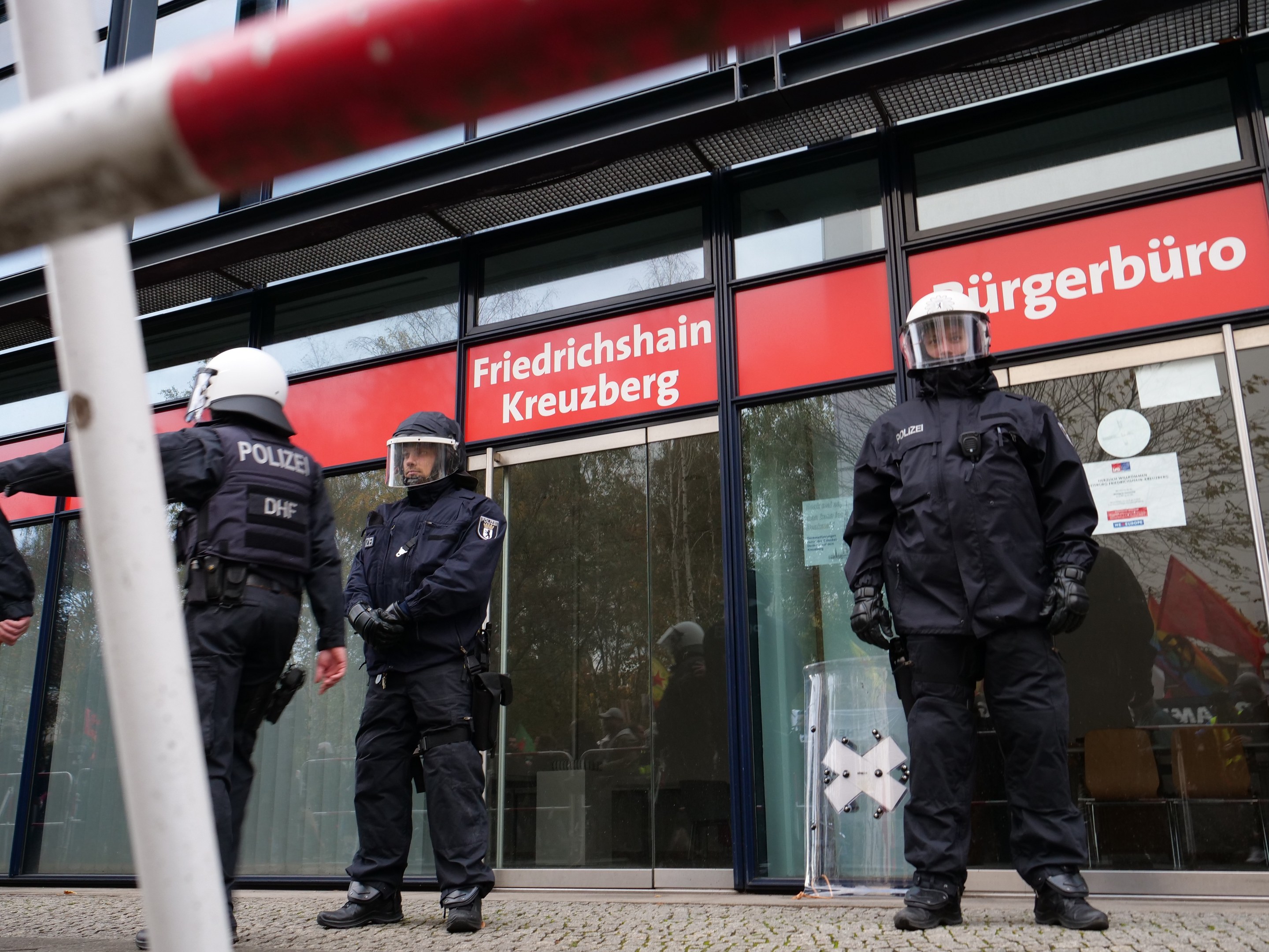 Polizisten in Uniform vor einem Glasfrontgebäude mit einer Stange links und Schildern mit der Aufschrift "Friedrichshain Kreuzberg" im Hintergrund.
