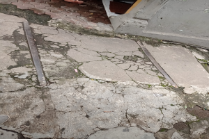 Eine Person steht auf einem gebrochenen Gehweg neben einem Gebäude, mit einem Metallobjekt auf dem Boden, einer Wand und einer Tür.