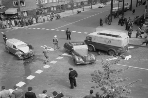 Schwarz-weiß-Szene eines Autounfalls an einer Straßenkreuzung mit mehreren Fahrzeugen, darunter ein Lieferwagen, und Menschen in der Nähe; Gebäude, Bäume, Pfosten und Texttafeln im Hintergrund.