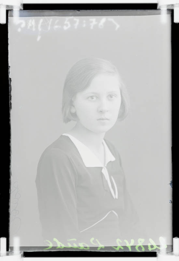 Ein junges Mädchen in Schuluniform steht vor einem dunklen Hintergrund, mit Text auf dem schwarz-weißen Foto aus den frühen 1900er Jahren.