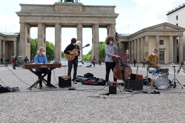 Eine Gruppe von Menschen, die Musik im Freien spielen, mit einem Gitarristen im Sitzen, einem anderen Musiker und einer maskierten Person, vor dem Hintergrund des Brandenburger Tors in Berlin.