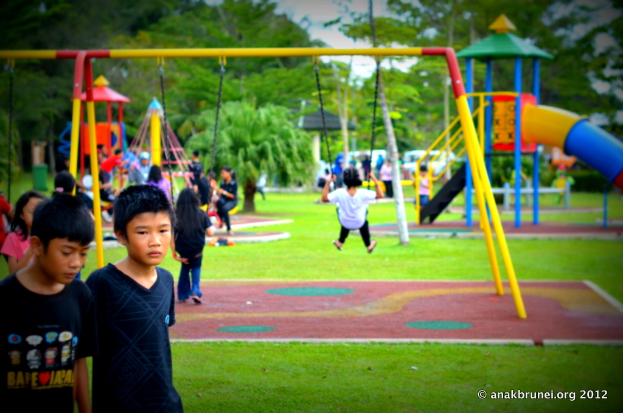Kinder beim Spielen auf Spielgeräten in einem Park mit Bäumen im Hintergrund.