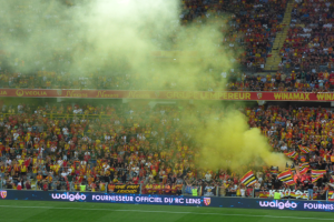 Ein Fußballstadion voller Menschen, einige halten Fahnen und Schilder, andere stehen oder sitzen, mit einer Rauchwolke in der Luft.