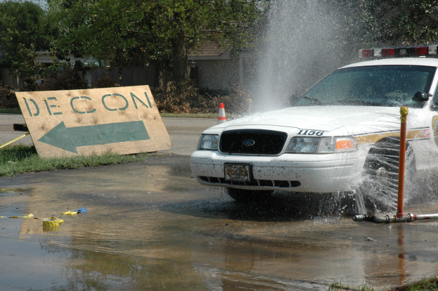 Ein Polizeiwagen fährt durch eine Pfütze, mit einer Tafel, Verkehrskegel und anderen Gegenständen am Boden, mit Bäumen, Gebäuden und einem klaren blauen Himmel im Hintergrund.