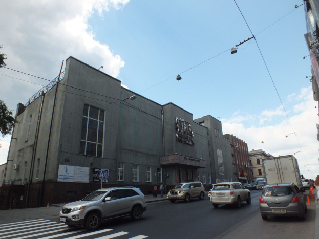 Stadtstraße mit fahrenden Autos, einer Zebrastreifen, Verkehrskegeln, Fußgängern auf dem Gehweg, Gebäuden, Laternen, Strommasten mit Kabeln, Bäumen und einem bewölkten Himmel.