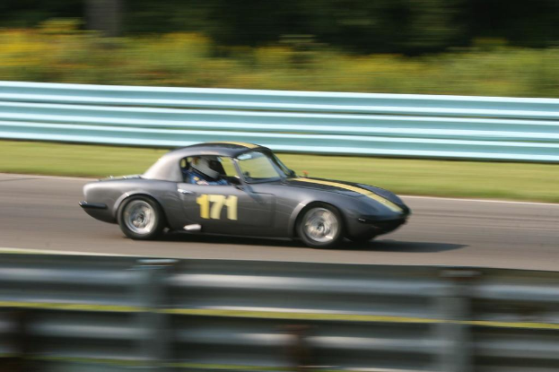 Ein Auto mit einem Helm tragenden Fahrer fährt auf einer Straße entlang, die von Barrieren gesäumt ist, mit Bäumen im Hintergrund.