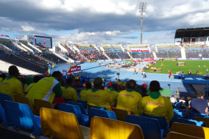 Eine Gruppe von Menschen schaut ein Fußballspiel in einem vollen Stadion mit Bannern, einem Bildschirm und einem bewölkten Himmel.