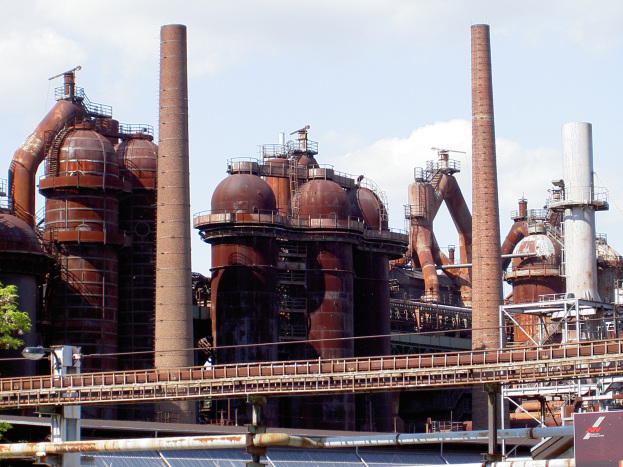 Ein großes Fabrikgebäude mit zahlreichen Rohren und Schornsteinen, einer Brücke, Polen, Lampen, Bäumen und einem bewölkten Himmel im Hintergrund.