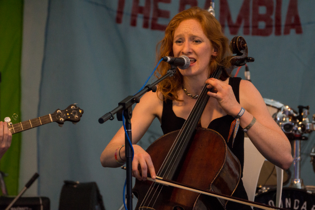 Eine Frau in einem schwarzen Kleid spielt Geige vor einem Mikrofon auf einem Ständer, mit einem Banner und einer Band im Hintergrund.