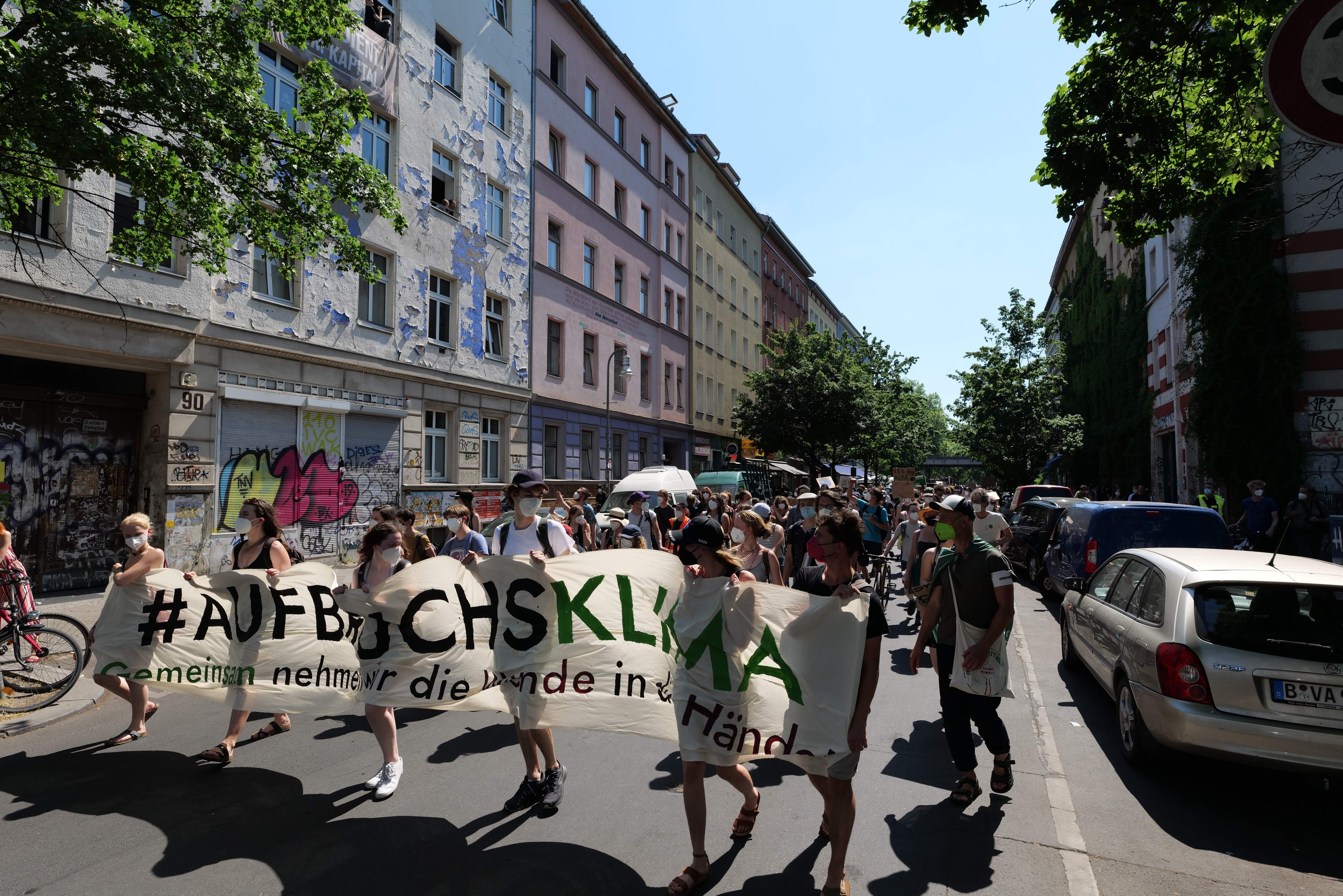Eine Gruppe von Menschen marschiert mit einem Transparent die Straße entlang, Autos parken rechts, ein Fahrradfahrer links, vor Gebäuden und Bäumen, während einer Demonstration in Berlin.