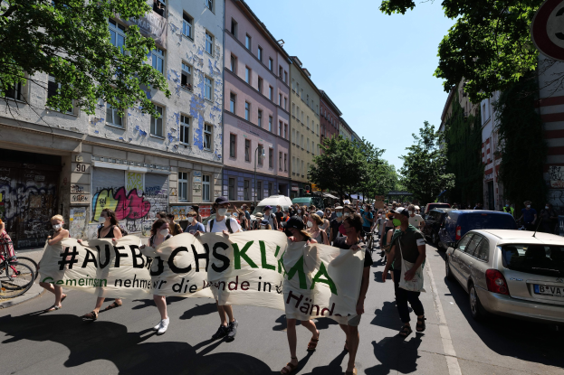 Eine Gruppe von Menschen marschiert mit einem Transparent die Straße entlang, Autos parken rechts, ein Fahrradfahrer links, vor Gebäuden und Bäumen, während einer Demonstration in Berlin.