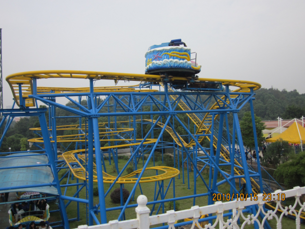 Achterbahn in einem Park mit Menschen, Zelten, Bäumen, einem Turm und einem bewölkten Himmel.