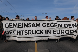 Eine Gruppe von Menschen steht auf der Straße und hält ein Schild hoch, auf dem "Gemeinsam gegen den Rechtsruck in Europa" steht, mit Straßenlaternen, Strommasten, Stromkabeln, Gebäuden und einem bewölkten Himmel im Hintergrund.