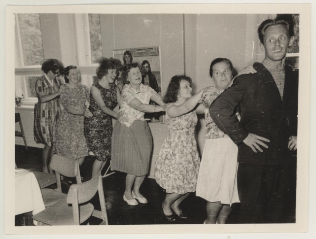 Eine Gruppe von Menschen, die in einem Raum mit Stühlen und einem mit einer weißen Decke bedeckten Tisch tanzen. An der Wand befindet sich ein Fotoраhmen und im Hintergrund sind Fenster zu sehen.