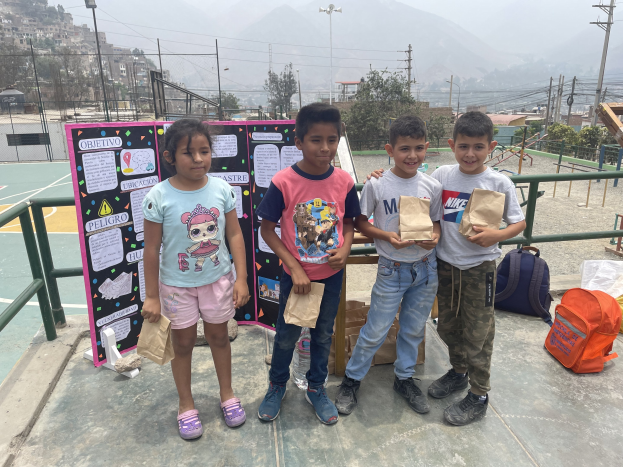 Gruppe von Kindern auf einem Basketballfeld, die Papiertüten halten und Gegenstände auf dem Boden rechts und eine Tafel mit Text links, vor einer Außenkulisse mit Zaun, Pfosten und Gebäuden.