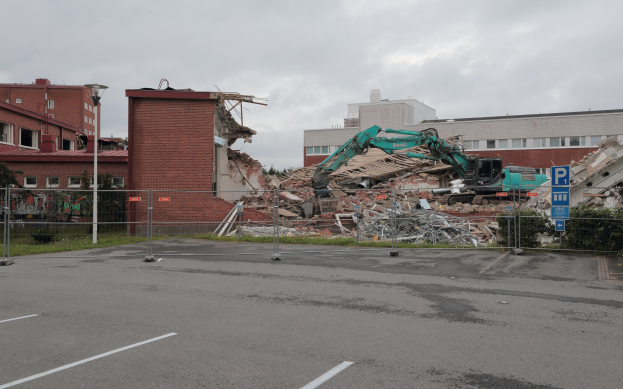 Großes Gebäude in einer Parkfläche mit einem Bagger, Metallstangen, Zaun, Schild, Pflanzen, Gras, Bäumen, benachbarten Gebäuden und einem bewölkten Himmel abgebrochen.