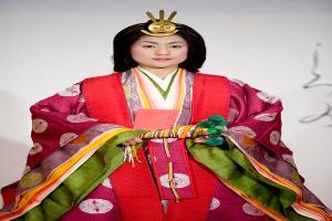Eine Frau in einem traditionellen japanischen Kimono, das ein leuchtend rotes und grünes Kleid mit kunstvollen Mustern zeigt, steht selbstbewusst auf einem Laufsteg, ihr Haar ist ordentlich zu einem Knoten gebunden.