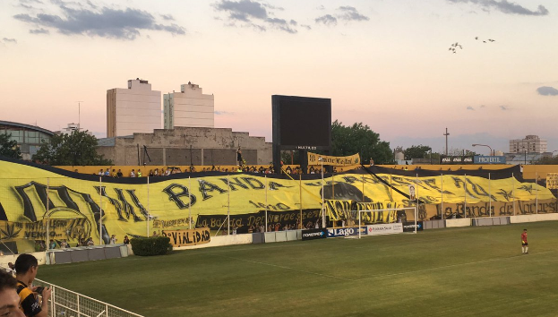 Ein Fußballfeld mit einer Gruppe von Menschen darauf, umgeben von einem Metallzaun, einem Torpfosten, einer Banner mit der Aufschrift "Boca Juniors vs Real Sociedad", einigen Pflanzen, einem Anzeigebildschirm, einigen Pfählen, der Rinde eines Baumes, einigen Bäumen, einigen Gebäuden und einem bewölkten Himmel.