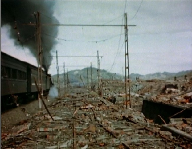 Ein Zug fährt durch ein beschädigten Gebiet mit Rauch, der aus ihm aufsteigt, umgeben von Strommästen und -drähten, während Hügel und ein klarer Himmel im Hintergrund zu sehen sind und zerstreute Trümmmer am Boden liegen.