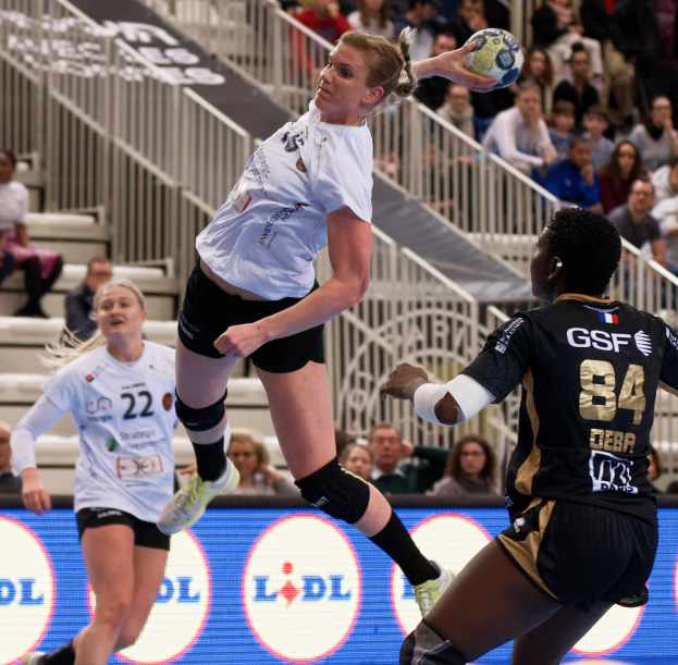 Eine Gruppe von Frauen beim Handballspielen auf einem Platz mit einem Ball in der Luft, Zuschauern auf Treppen und Geländern im Hintergrund und einem Schild mit der Aufschrift "Handball-Weltmeisterschaft 2019 - Tag 2".