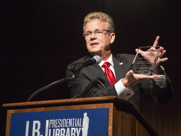 Ein Mann im Anzug und Krawatte steht an einem Rednerpult und hält eine Auszeichnung in der Hand, mit einem Mikrofon und einer Tafel am Rednerpult.