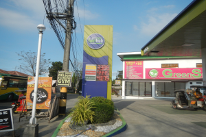Tankstellen-Schild mit der Aufschrift 'Green's' mit Fahrzeugen, Pflanzen, Infrastruktur und Gebäuden im Vordergrund und Bäumen und Wolken im Hintergrund.