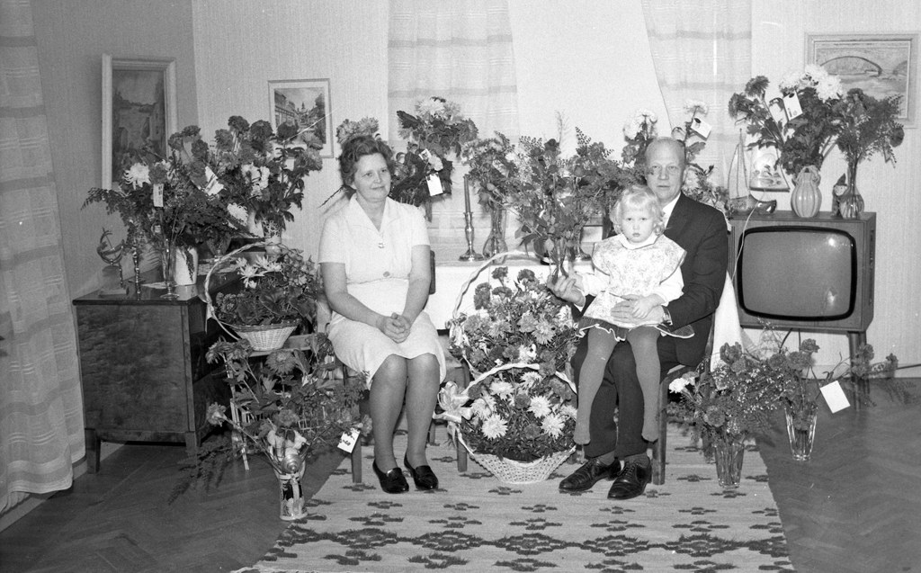 Schwarzes und weißes Foto einer Familie aus drei Personen - einem Mann, einer Frau und einem Kind - auf einem Sofa sitzend mit Blumenvasen auf nahen Tischen und einem Teppich auf dem Boden, gerahmte Fotos an der Wand und Vorhänge im Hintergrund.