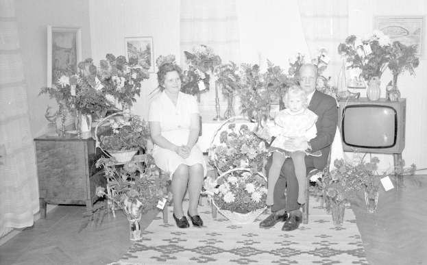 Schwarzes und weißes Foto einer Familie aus drei Personen - einem Mann, einer Frau und einem Kind - auf einem Sofa sitzend mit Blumenvasen auf nahen Tischen und einem Teppich auf dem Boden, gerahmte Fotos an der Wand und Vorhänge im Hintergrund.