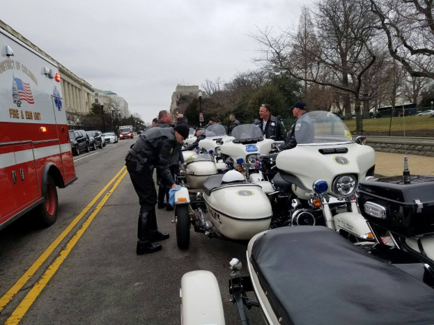 Eine Gruppe von Polizeibeamten neben Motorrädern am Straßenrand stehend, mit Fahrzeugen wie einem Löschfahrzeug, Bäumen, Gebäuden und einem klaren blauen Himmel im Hintergrund.