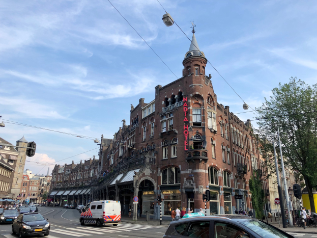 Eine belebte Stadtstraße in Amsterdam mit Autos, Fußgängern, hohen Gebäuden, Bäumen und Ampeln unter einem bewölkten Himmel.