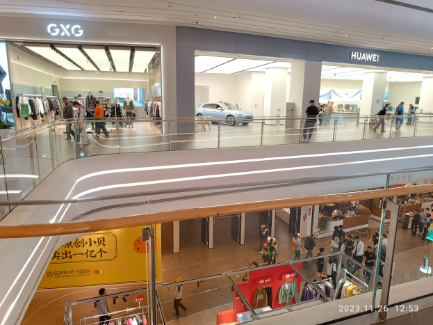 Ein Einkaufszentrum-Innenraum mit Menschen, die gehen, Glasgeländern, Bekleidungsauslagen, Informationsschildern, Säulen, Deckenleuchten und einem geparkten Auto im Hintergrund, mit einem GXG- und Huawei-Partnerschafts-Wasserzeichen.