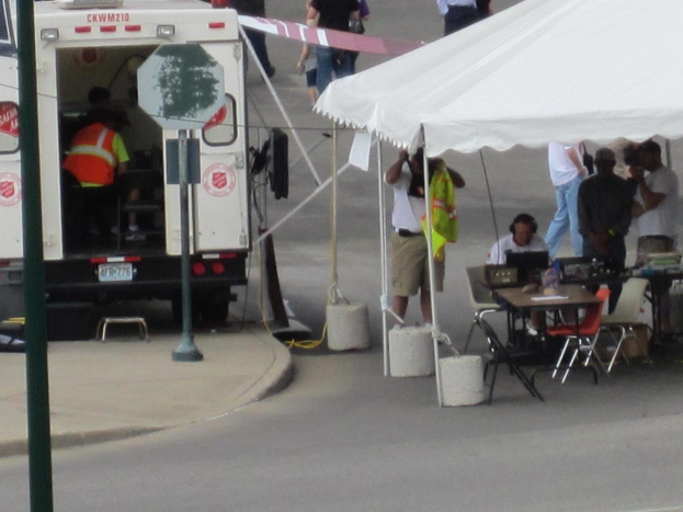 Ein Krankenwagen steht auf der linken Seite einer StraÃŸe, wÃ¤hrend auf der rechten Seite ein Zelt viele Menschen schÃ¼tzt, mit einem Tisch und einem Stuhl in der NÃ¤he.