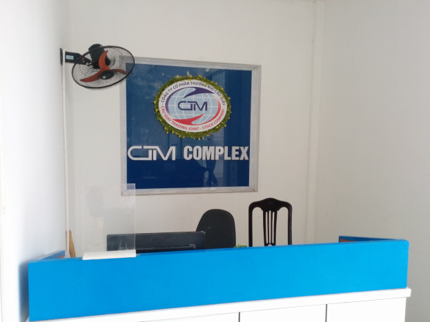 Ein blauer und weißer Empfangstisch mit einem Computer, umgeben von Stühlen und einem Tischventilator, mit einer Wandtafel, die Text und ein Logo anzeigt, das darauf hinweist, dass es das Büro von GM Trading Joint in Ho Chi Minh Stadt ist.