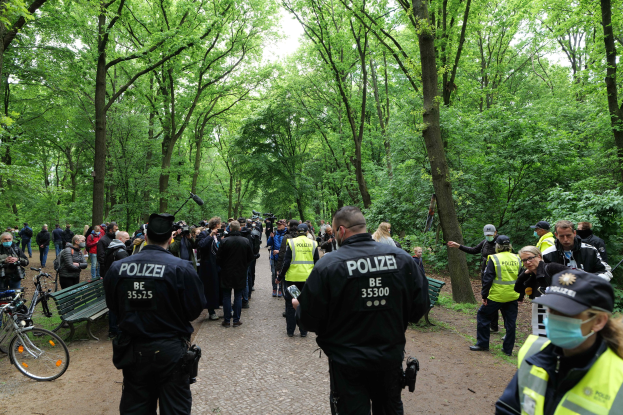 Polizeibeamte vor einer Menschenmenge bei einer Outdoor-Veranstaltung, mit Fahrrädern und einer Bank im Vordergrund und Bäumen im Hintergrund.