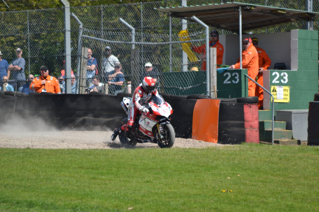 Eine Person in Helm und Handschuhen fährt ein Motorrad über ein grasbewachsenes Feld, mit Zuschauern in Mützen und Schutzbrillen, Maschendrahtzaun, Treppe mit Geländer und einem nummerierten Schuppen im Hintergrund.