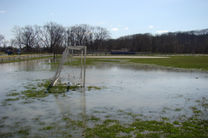 Ein Fussballtor steht auf einem überfluteten Feld, umgeben von Gras, einem Zaun, Bäumen, Häusern und unter einem bewölkten Himmel.