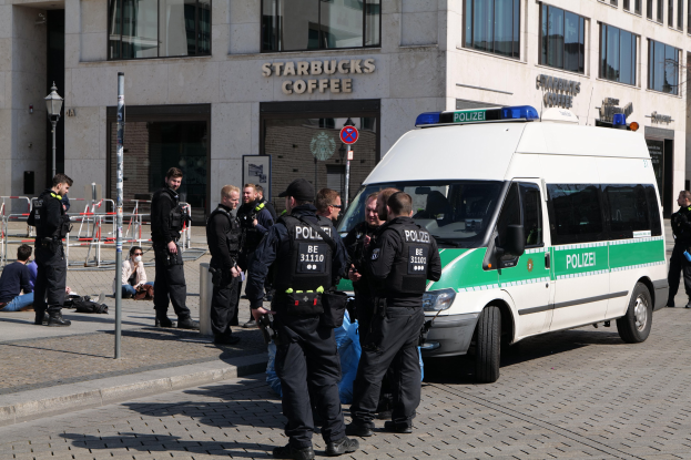Eine Gruppe von Polizisten steht vor einem Starbucks-Café, rechts davon ein Van und links ein paar Menschen; im Hintergrund sind ein Gebäude, ein Schild, ein Lichtmast und ein Zaun zu sehen.