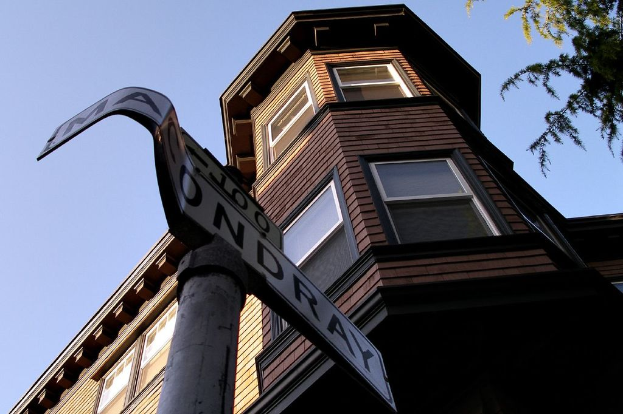 Ein Pfahl mit Text vorne, ein Gebäude in der Mitte und Blätter auf der rechten Seite.